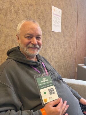 Man, indoors, with grey hair smiling and holding up name tag