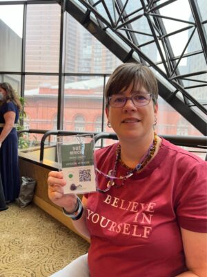 Woman, indoors, with red t-shirt smiling and holding up name tag