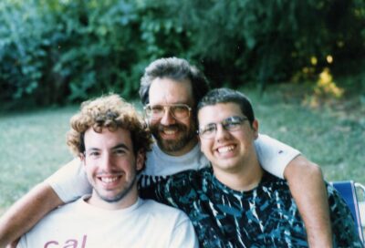 Three smiling men in the late 90's outdoors. Father with arms around 2 sons