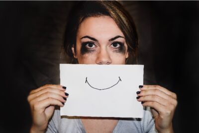 A person with smudged eyes holding a piece of paper with a drawn smile over mouth