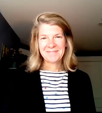 Smiling physician standing looking into video camera, blond hair, stripe shirt, dark sweater