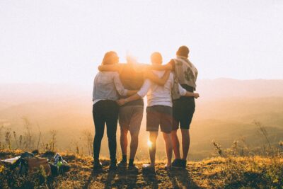 Photo looking at the backs of 4 people hugging with sun directly in front on a hill's edge