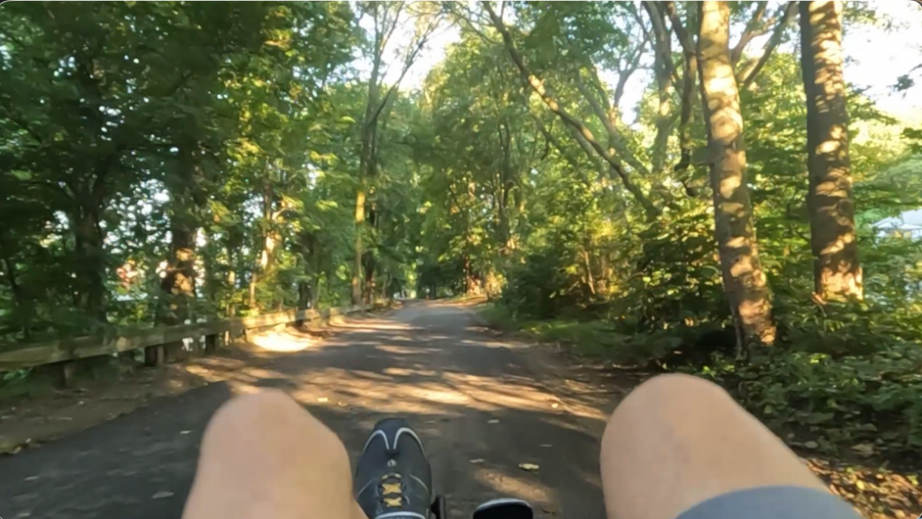 First-person view along a tree-lined park path, with legs and sneakers in the foreground on a bike ride.