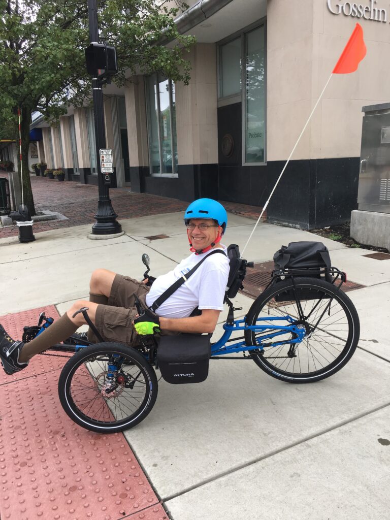 Smiling man in a blue helmet sits on a blue recumbent tricycle with side pannier bags on a city sidewalk.